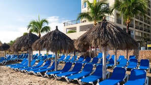 Una playa con filas de sillas azules y sombrillas de paja, una palmera y un edificio al fondo.