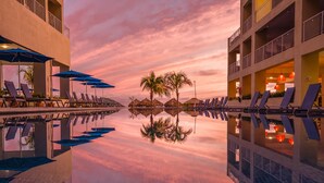 Zona de piscina de hotel con sillas de descanso y sombrillas, palmeras y un reflejo en el agua.
