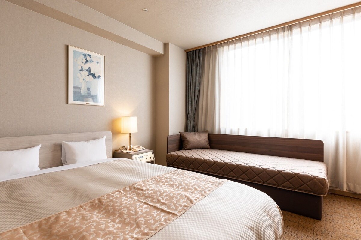 Une chambre d’hôtel avec un grand lit, un lit plus petit, une table de chevet avec une lampe et une fenêtre avec des rideaux.