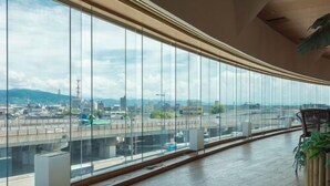 A modern interior with large windows offering a city view, a wooden floor, and a seating area with a plant. - Tennen Onsen Relax Park Terume Kanazawa (Kanazawa)