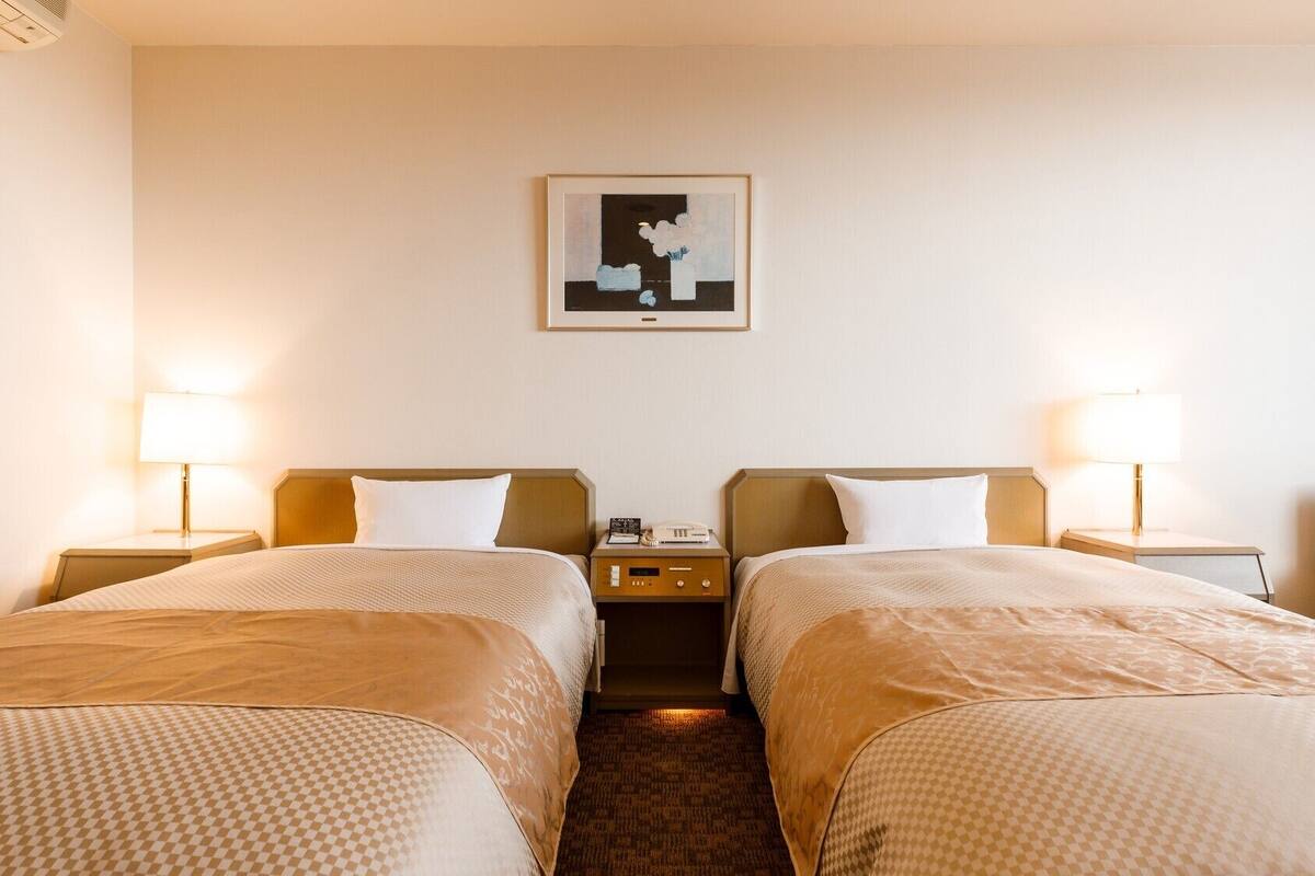 A hotel room with two beds, bedside lamps, a nightstand with a phone, and a framed picture on the wall.
