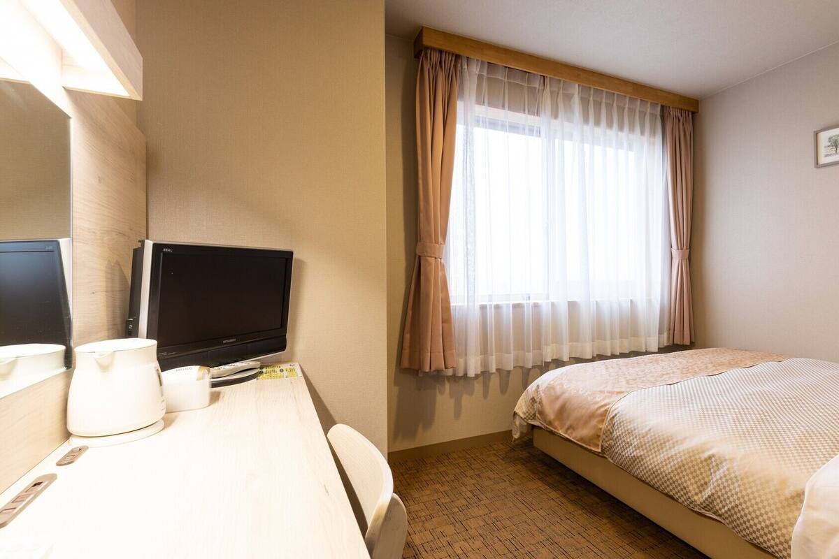 A hotel room with a bed, a desk with a television, a coffee maker, and a window with sheer curtains.