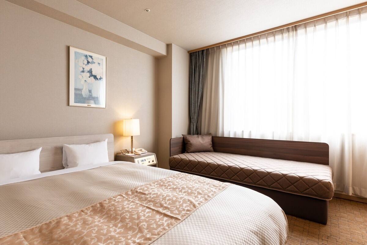 A hotel room with a large bed, a smaller bed, a bedside table with a lamp, and a window with curtains.