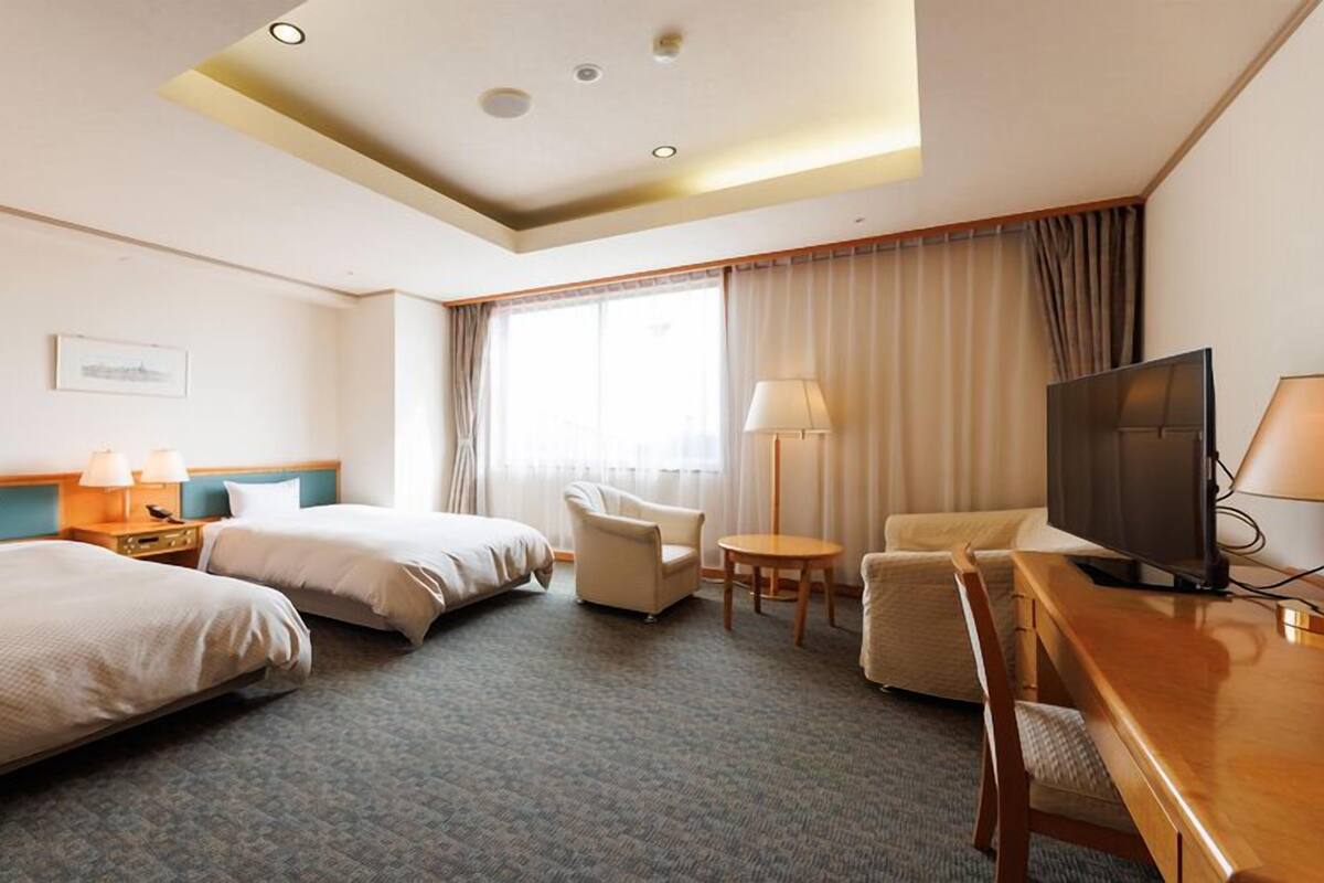 A hotel room with two beds, a desk with a TV, a chair, a lamp, and a window with curtains.