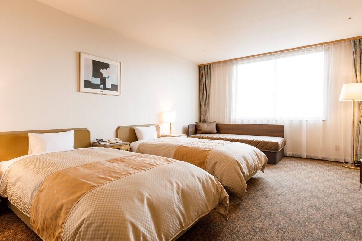A hotel room with two beds, a bedside table with a lamp, a large window with curtains, and a framed picture on the wall.