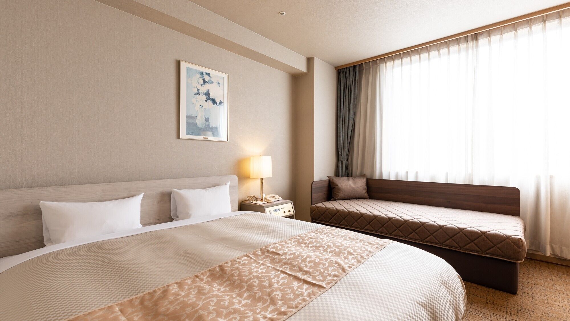 A hotel room with a large bed, a smaller bed, a bedside table with a lamp, a framed picture on the wall, and a window with curtains.