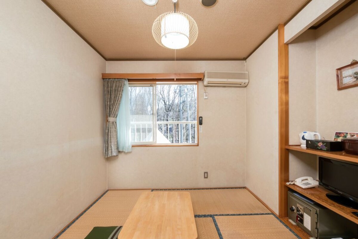 Ein Zimmer mit Tatami-Böden, einem Holztisch, einem Fernseher und einem Fenster mit Vorhängen.