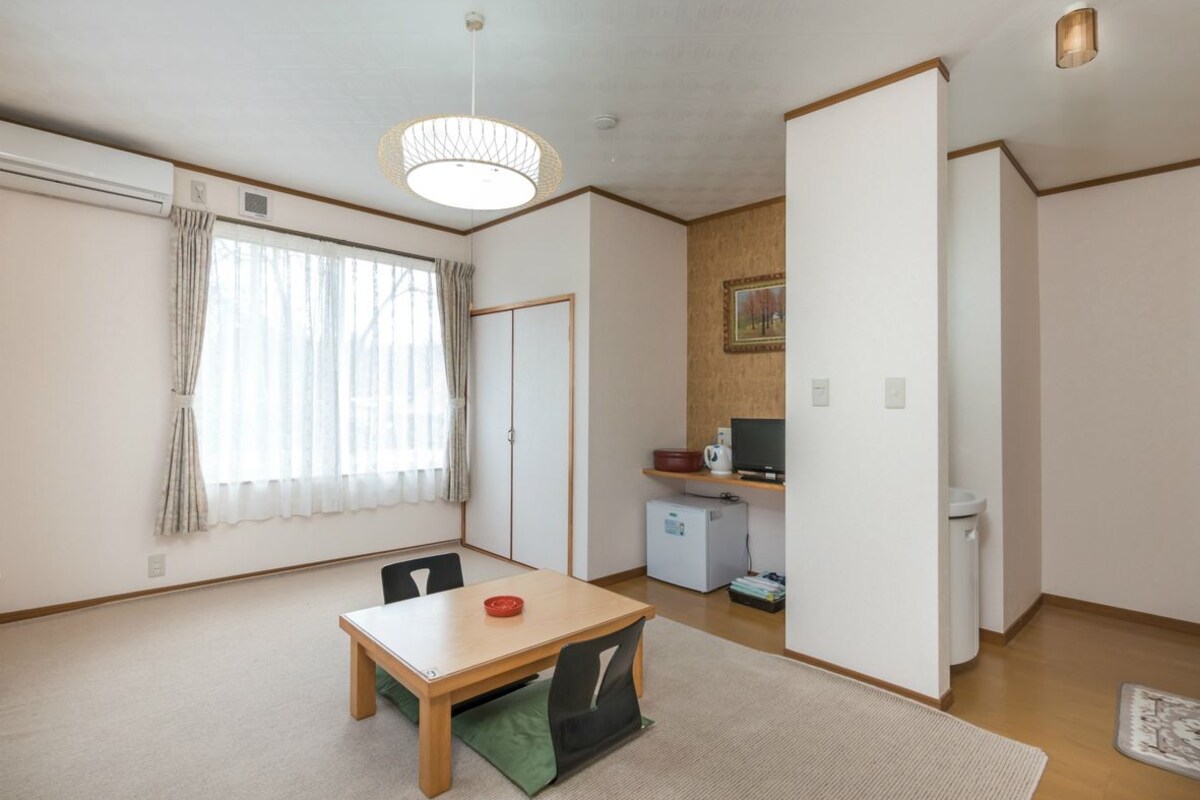 Ein Zimmer mit Esstisch, Stühlen, Fernseher, Kühlschrank und einer kleinen Küchenzeile.