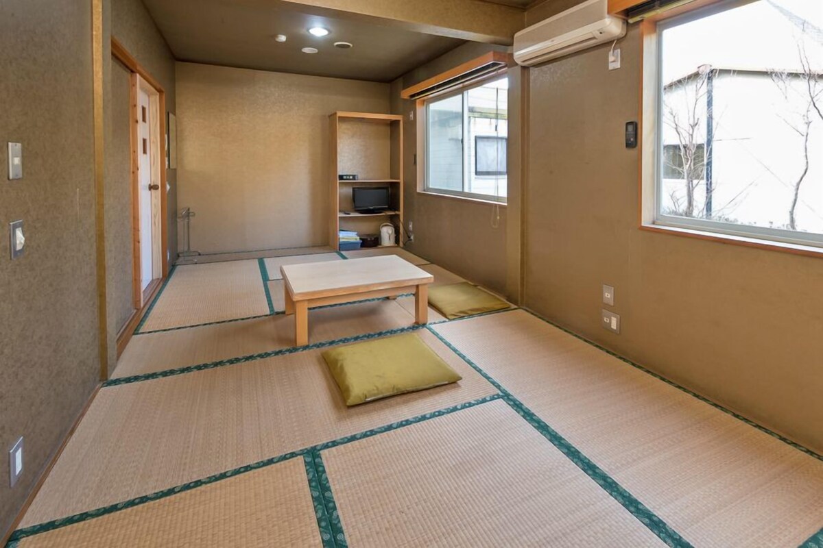 Ein Zimmer mit Tatami-Boden, einem Futon-Bett, einem Fernseher und einem Fenster mit Blick auf Bäume.