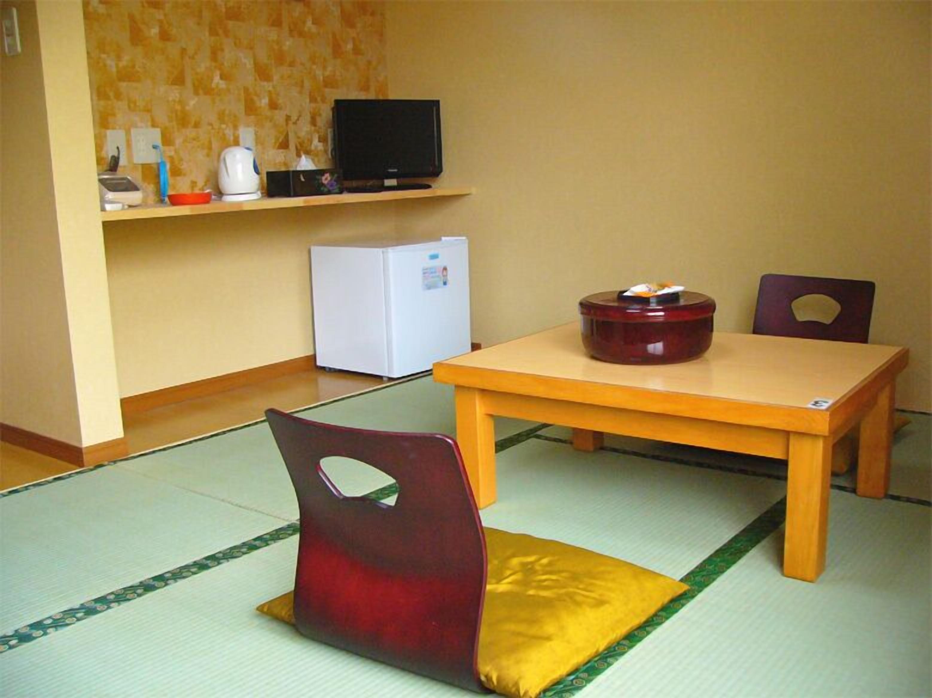 Una habitació amb sòl de tatami, taula de fusta, televisió sobre una estantalla, nevera i una cadira amb l'esquena orificària.