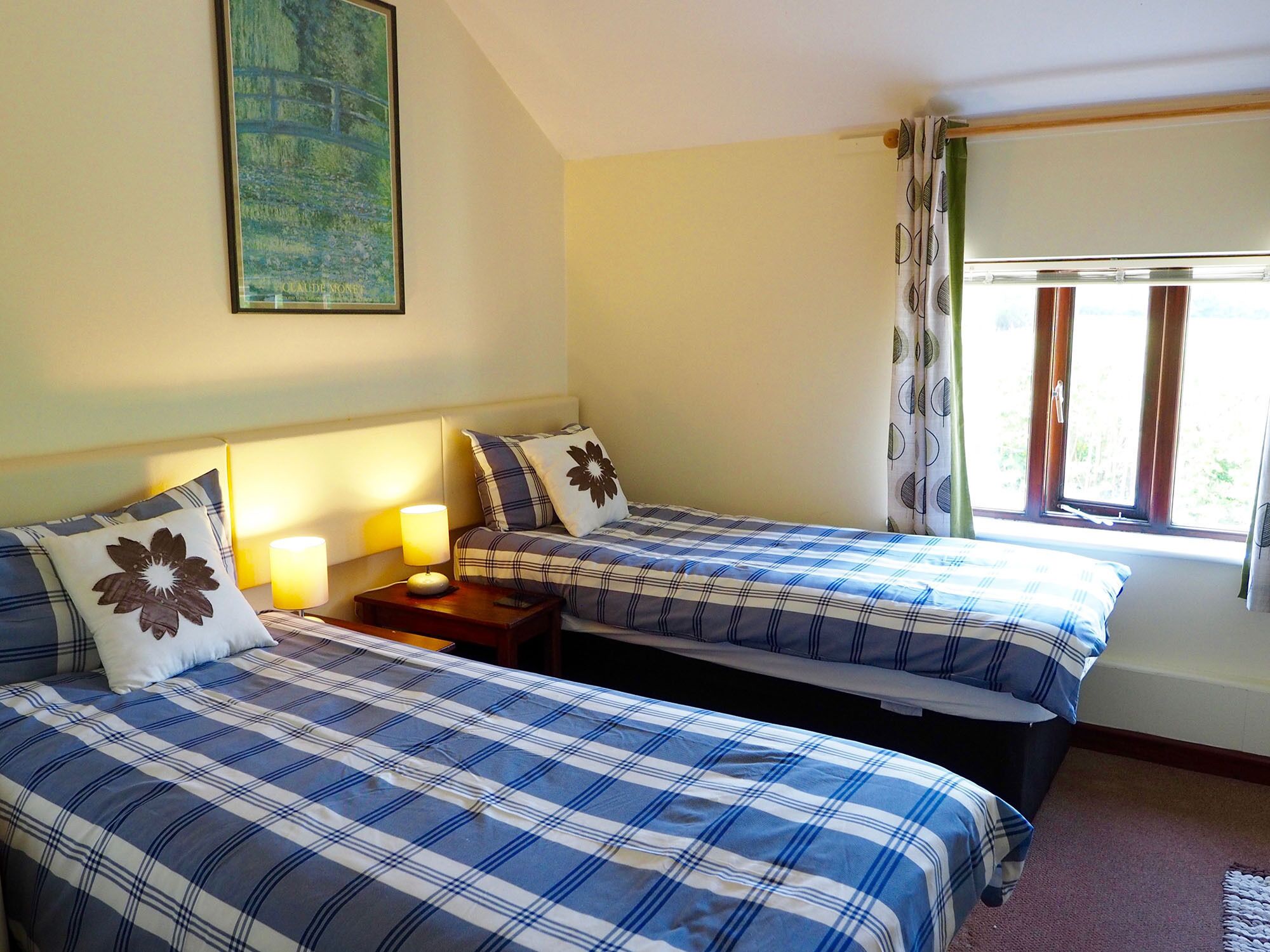 A bedroom with two beds, each with blue and white checkered bedding, a bedside table with a lamp, and a window with curtains.