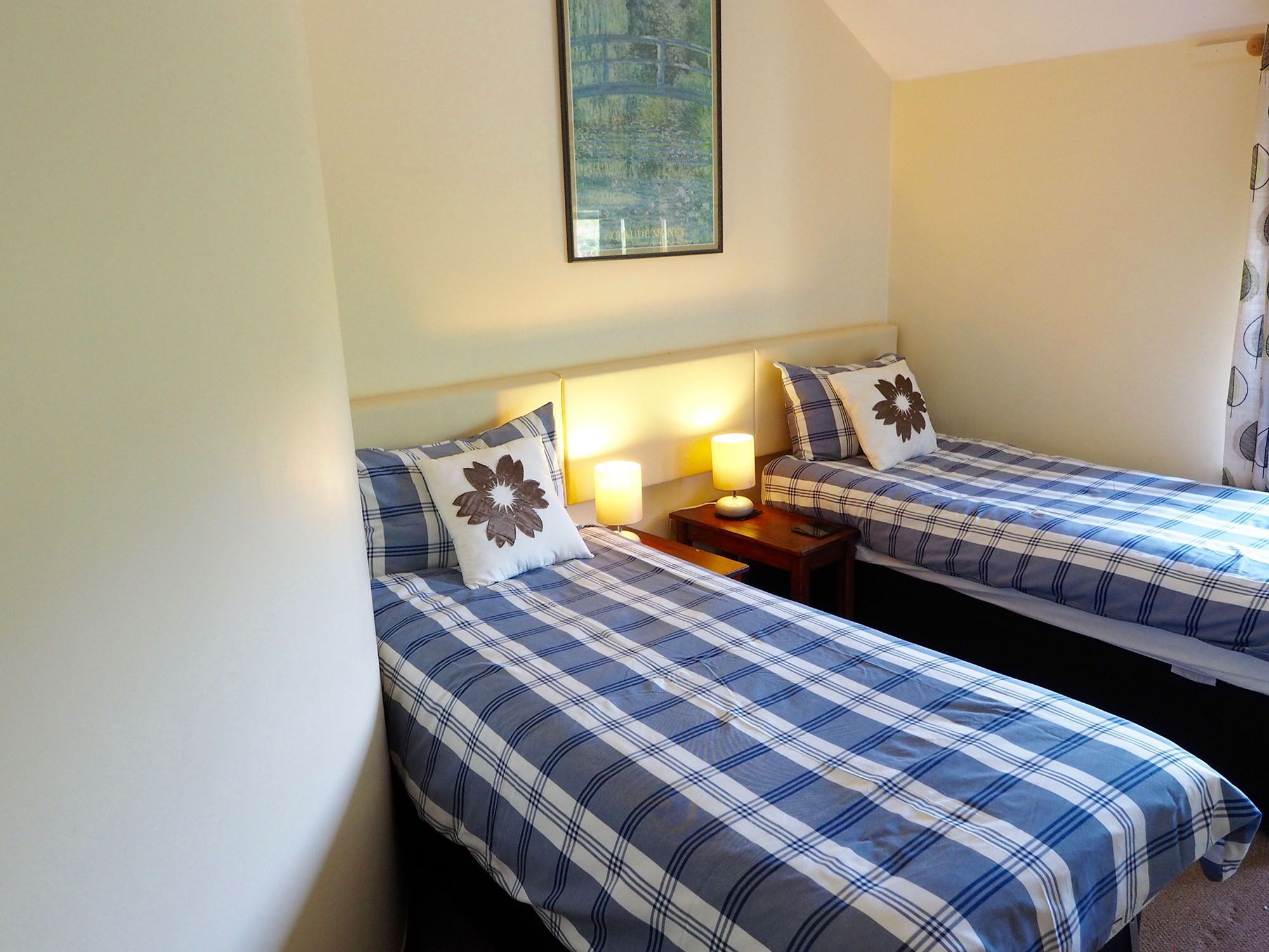 A room with two beds, each with a blue and white checkered bedspread and a decorative pillow. There is a nightstand with a lamp between the beds.