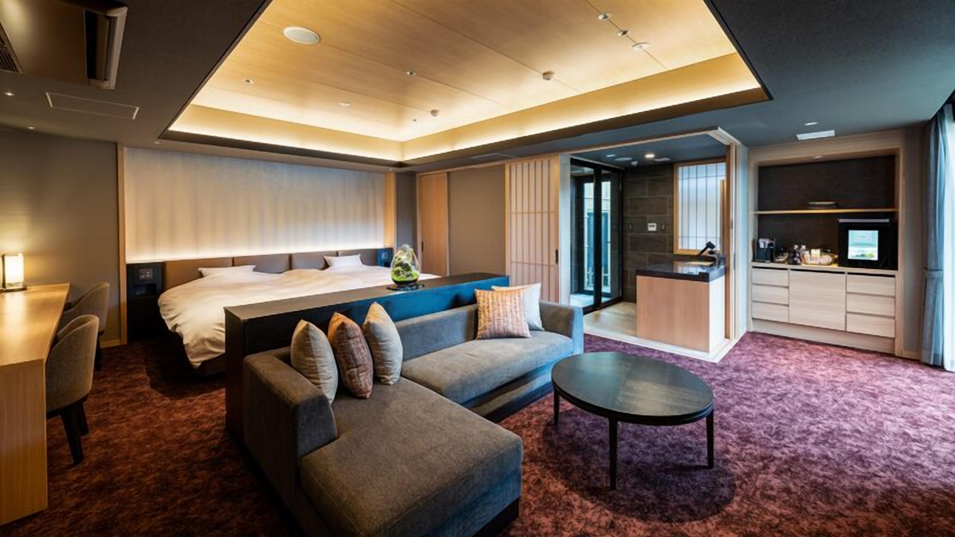 A modern hotel room with a bed, sofa, desk, and kitchenette.