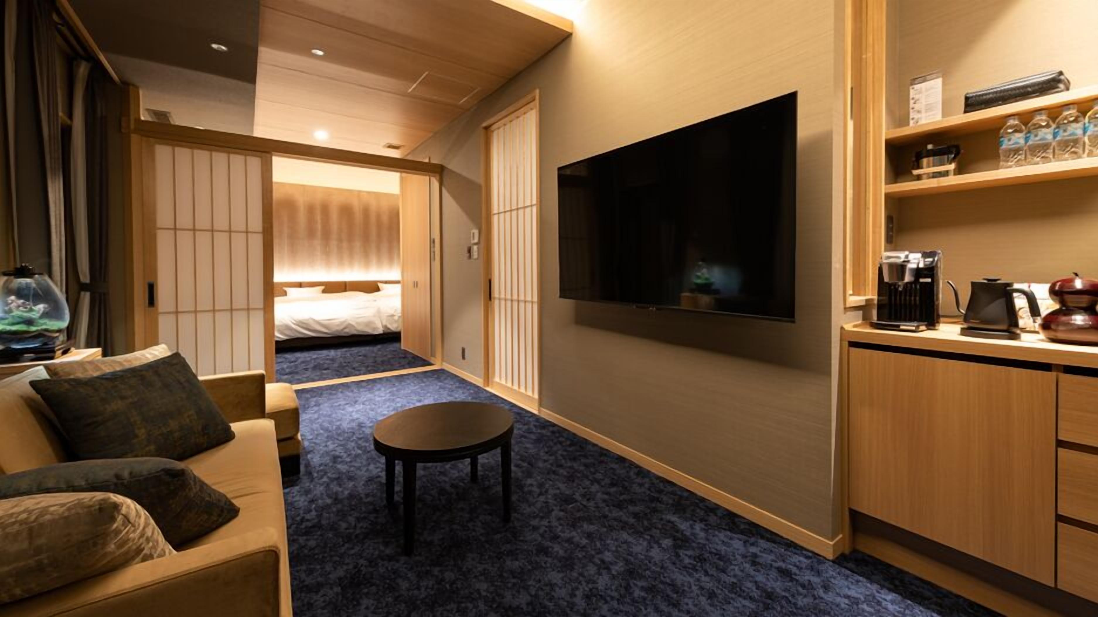 A hotel room with a bed, a TV, a small table, a sofa, and a kitchenette.