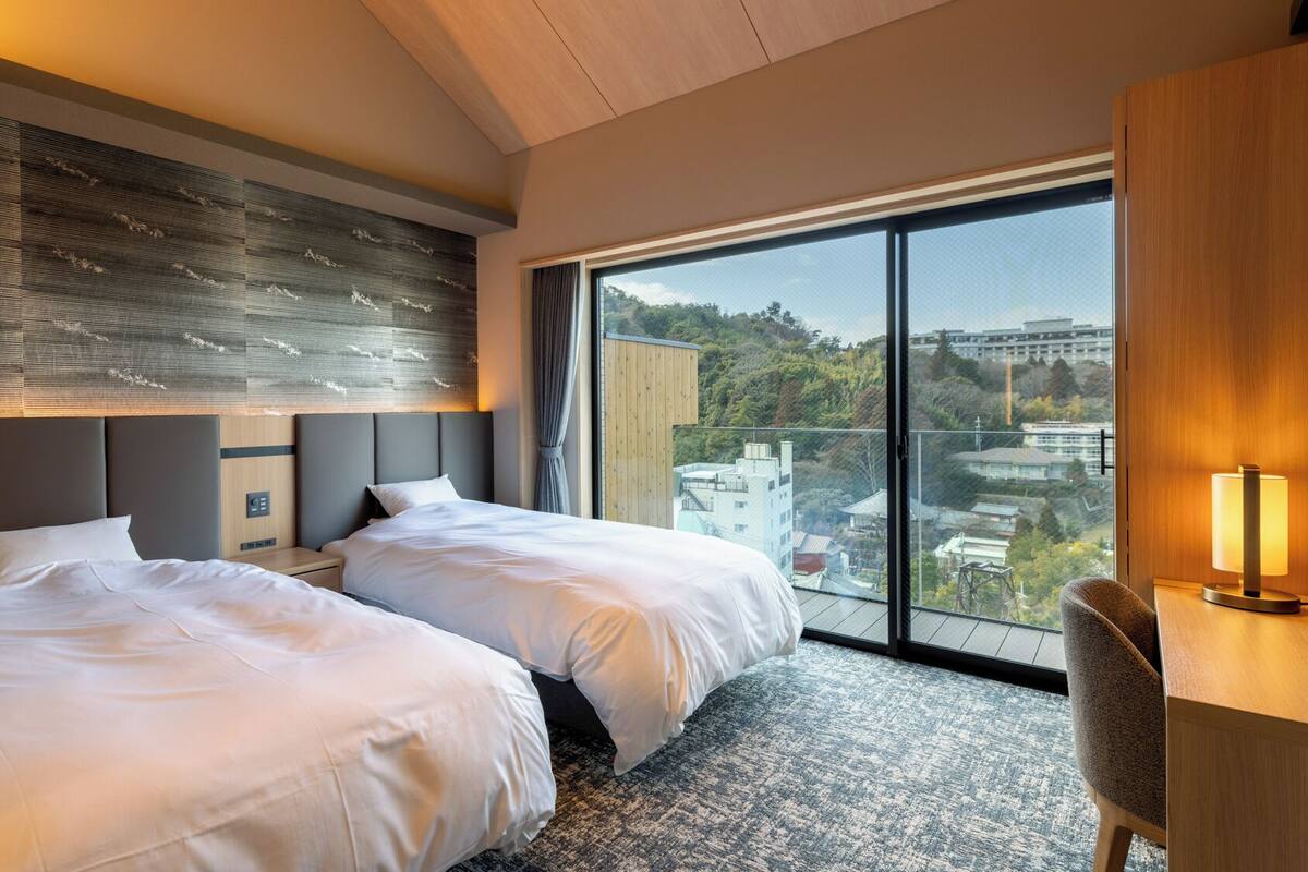 Habitación de hotel con dos camas, un ventanal amplio con vistas a la ciudad, un escritorio con lámpara y un cabecero moderno.