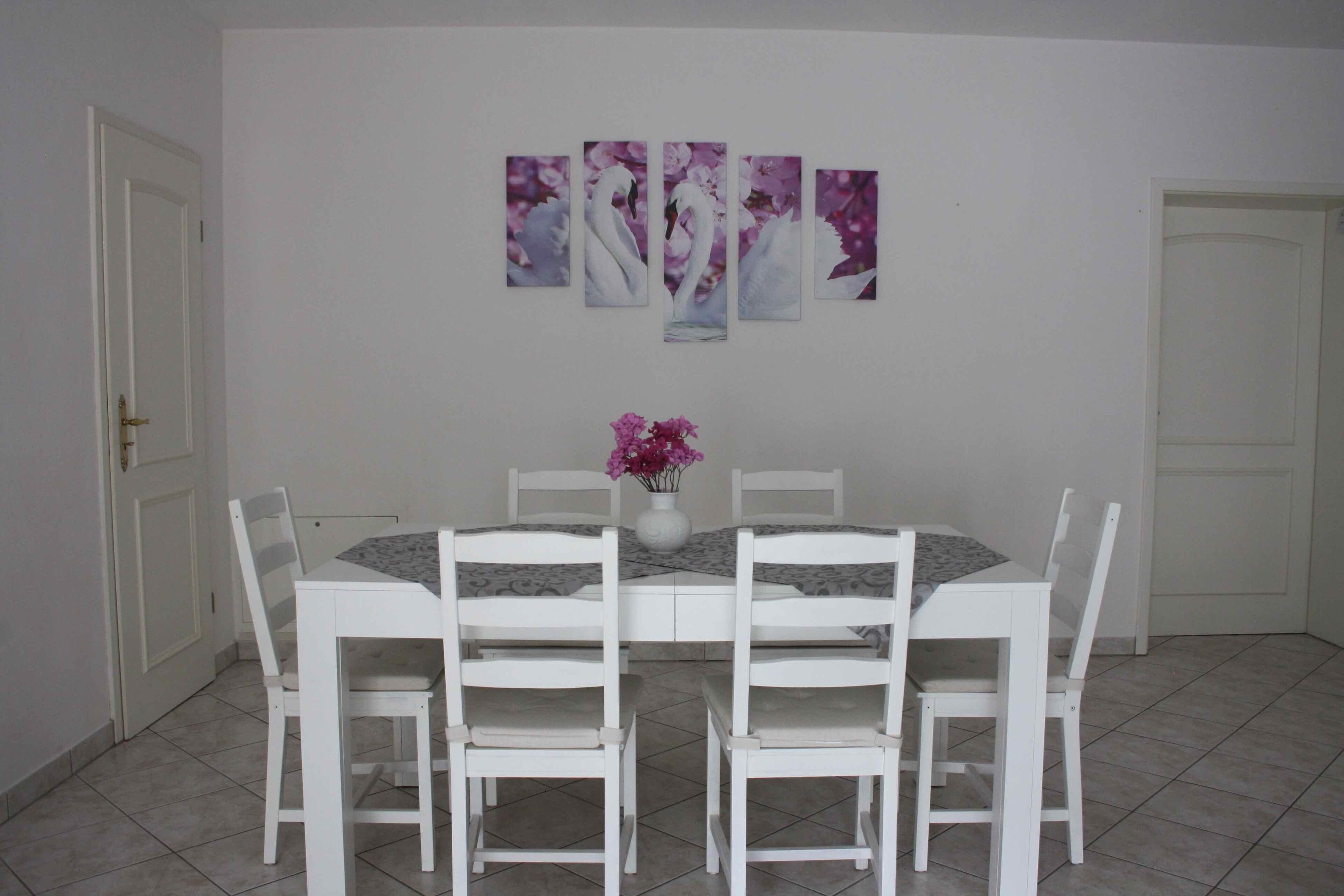 Un comedor con una mesa y sillas blancas, un jarrón con flores y una pared decorada con cuatro cuadros.
