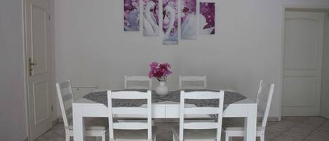 Un comedor con una mesa y sillas blancas, un jarrón con flores y una pared decorada con cuatro cuadros.