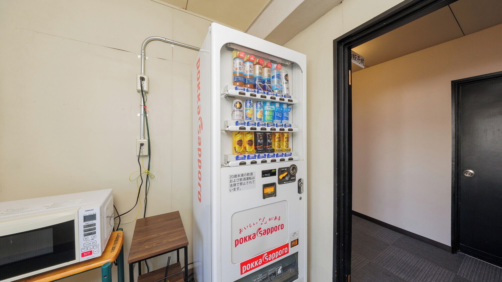 A vending machine with various beverages and snacks, a microwave, and a small wooden table.