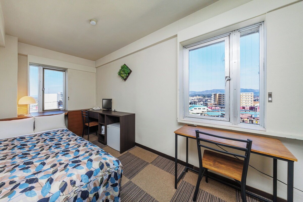 Ein Hotelzimmer mit Bett, Schreibtisch, Stuhl und großen Fenstern mit Blick auf die Stadt.