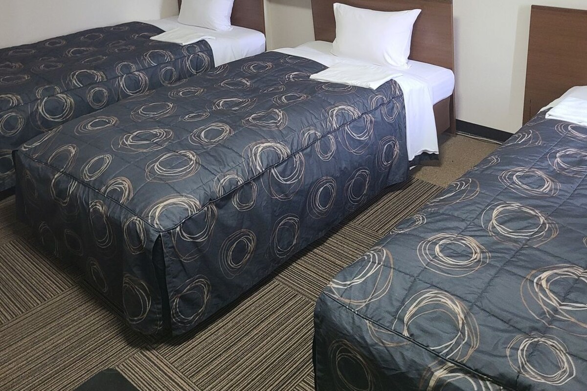 Ein Hotelzimmer mit drei Betten, jedes mit einer dunkelblauen Bettdecke mit kreisförmigen Mustern. Der Raum hat einen Teppichboden und ein gerahmtes Bild an der Wand.