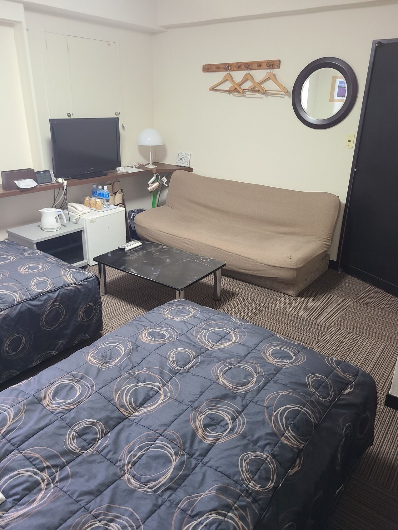 A hotel room with a sofa, two beds with patterned bedding, a small table, a TV, and a mirror on the wall.