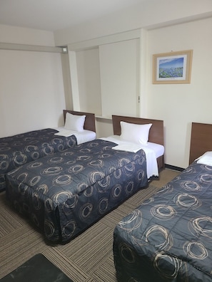 A hotel room with three beds, each with a dark blue bedspread featuring circular patterns. The room has a carpeted floor and a framed picture on the wall.