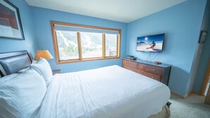 A bedroom with a large bed, a dresser, a TV, and a window with a mountain view.