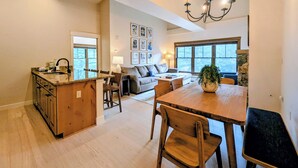 Une cuisine spacieuse avec un îlot central, un coin repas avec une table et des chaises en bois, un salon avec un canapé et une grande fenêtre, ainsi qu’un lustre.