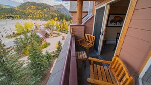 A wooden balcony with two wooden chairs and a table, overlooking a parking area and a mountain landscape with autumn-colored trees. - Summer Getaway | Hot Tub + Outdoor Fun (Keystone)