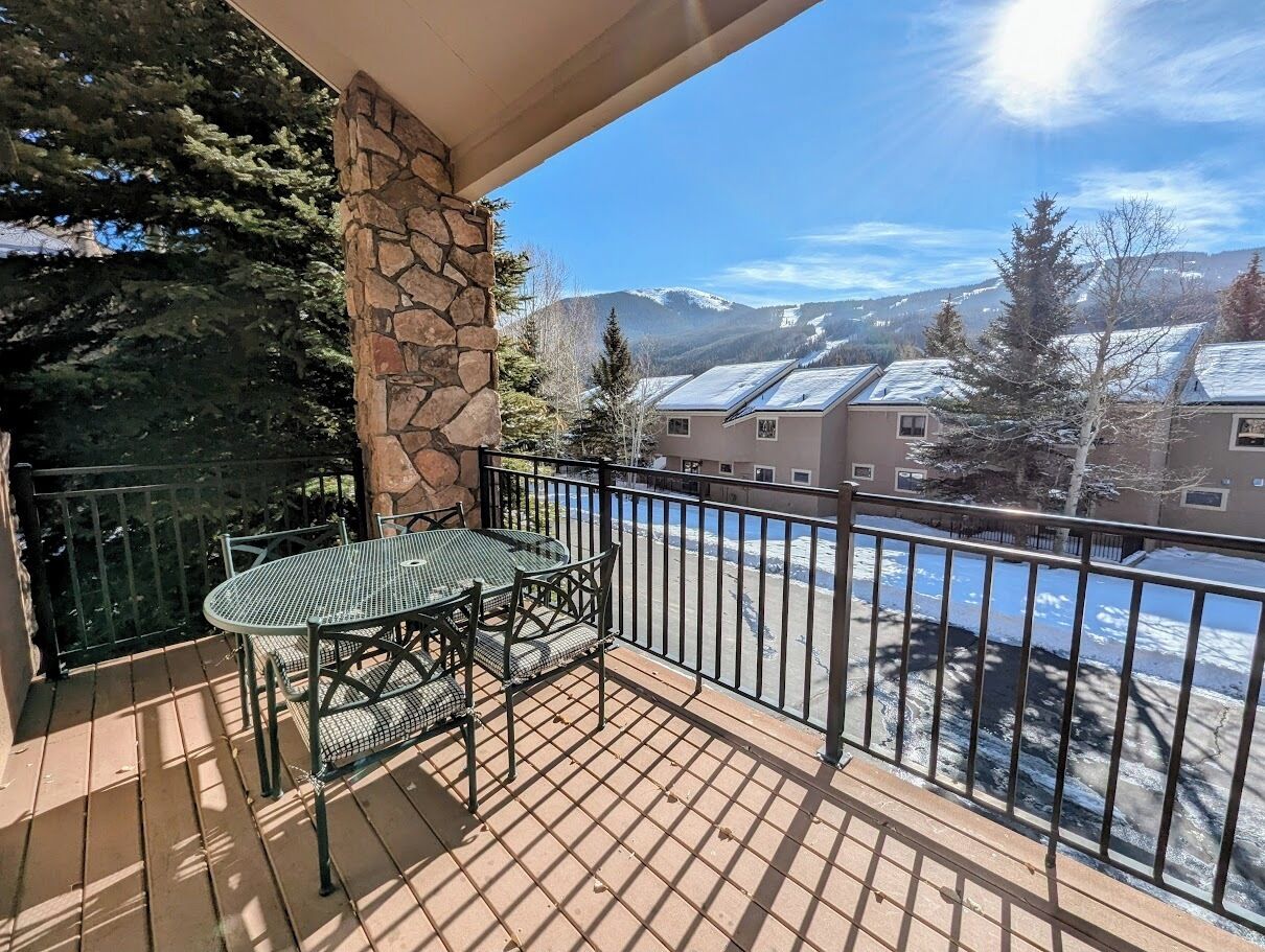 A balcony with a stone pillar, a metal table and chairs, and a view of snow-covered buildings and mountains.