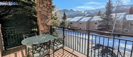 A balcony with a stone pillar, a metal table and chairs, and a view of snow-covered buildings and mountains.