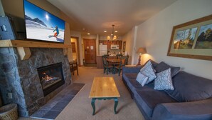 A living room with a stone fireplace, a television mounted above it, a wooden coffee table, a blue sofa with patterned cushions, and a dining area in the background.