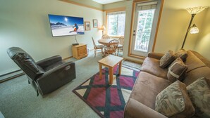 A living room with a leather sofa, a wooden coffee table, a TV mounted on the wall, and a window with a view of trees. - Summer Mountain Studio | Balcony & Hot Tub (Keystone)