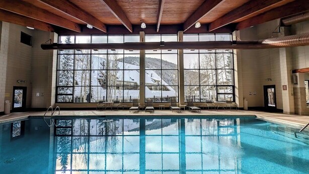 An indoor swimming pool with a view of snow-covered trees and buildings through large windows. - Cozy Condo w/Wood Fireplace & Indoor Pool/Hot Tubs (Keystone)