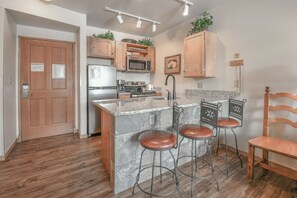Una cocina con armarios de madera, un refrigerador de acero inoxidable, un microondas y una estufa. Hay dos taburetes de bar y una silla de madera.