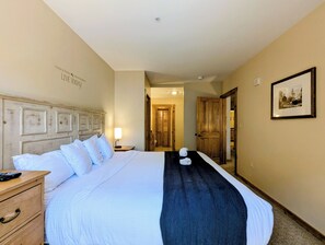 Une chambre d’hôtel avec un grand lit, une tête de lit en bois, une table de chevet avec une lampe, un tableau encadré au mur et une porte donnant sur un couloir.