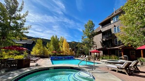 A swimming pool with a hot tub, lounge chairs, and umbrellas, surrounded by trees and a building with a balcony. - Corner Unit | Hot Tub & Village Summer Fun (Keystone)