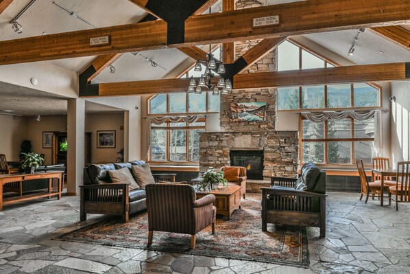 Cozy seating areas surround a central coffee table, with a stone fireplace and large windows offering views of the surrounding landscape.