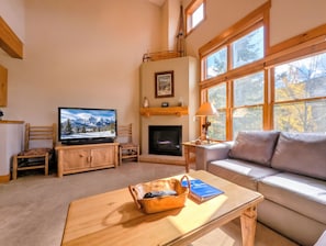A cozy living room with a television, fireplace, sofa, and a window with a view of trees.