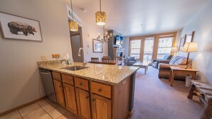 Cocina con encimeras de granito, armarios de madera y lavavajillas integrado. Hay un comedor con mesa y sillas, y una sala de estar con sofá y televisor.