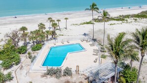 Ein Pool mit ungewöhnlicher L-Form, umgeben von einem Sandstrand und Palmen, mit freiem Blick auf den Ozean im Hintergrund.