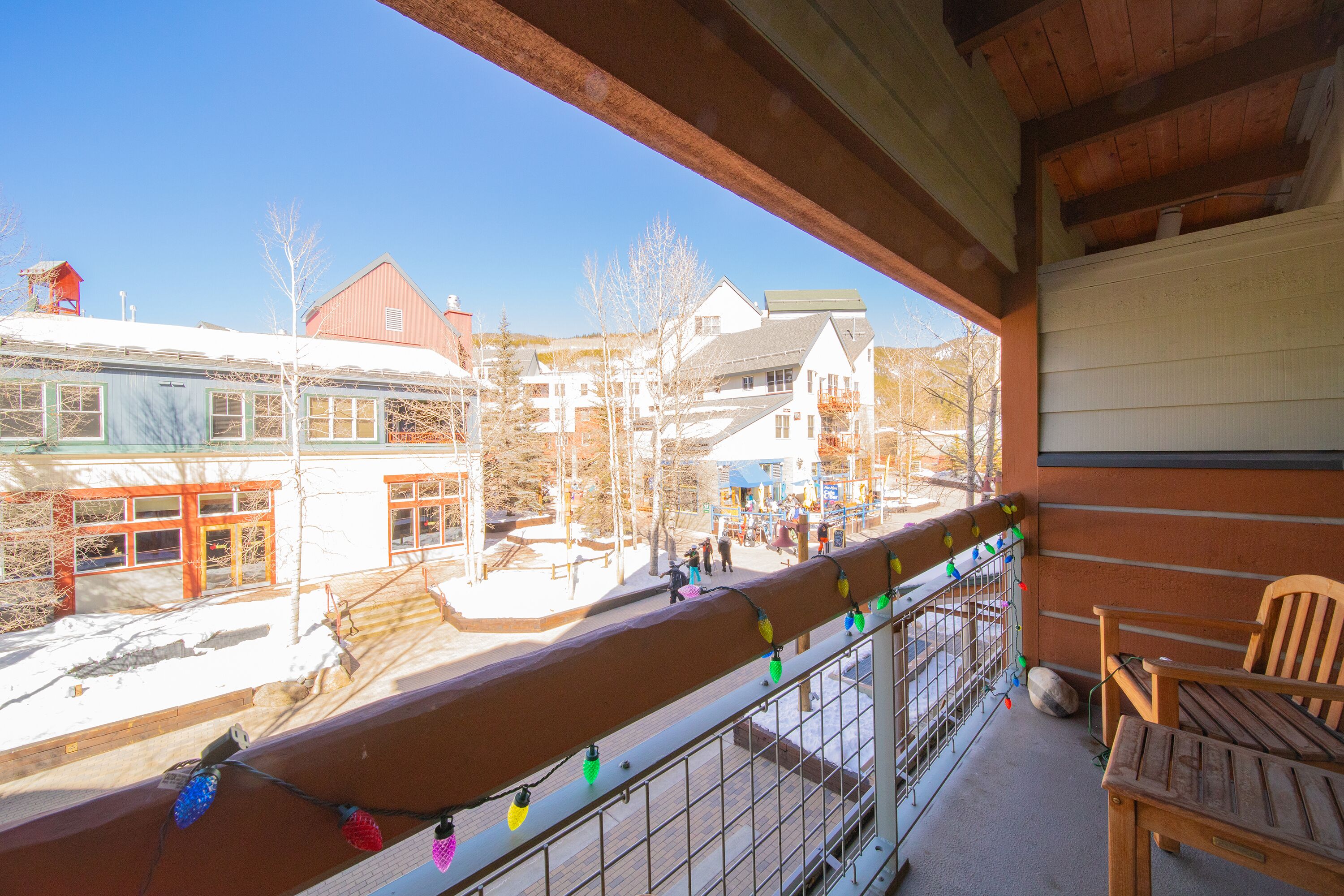 A balcony with a wooden chair, a railing with colorful lights, and a view of buildings and a playground.