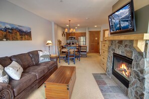 A living room with a brown leather sofa, a wooden coffee table, a stone fireplace, and a mounted TV. - Walk to River Run, Shops, Outdoor Pool/Hot Tub, (Keystone)