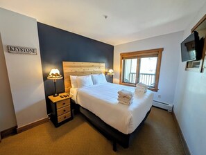 A neatly made bed with white linens and a wooden headboard, a nightstand with a lamp, and a window with blinds. - Walk to River Run, Shops, Outdoor Pool/Hot Tub, (Keystone)