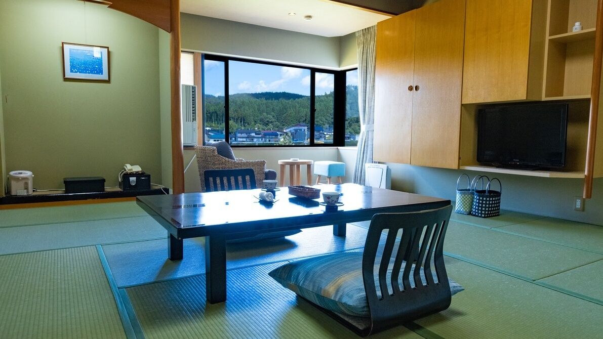 Una habitación con esteras de tatami, una mesa baja con sillas, un televisor y vistas a una zona boscosa.
