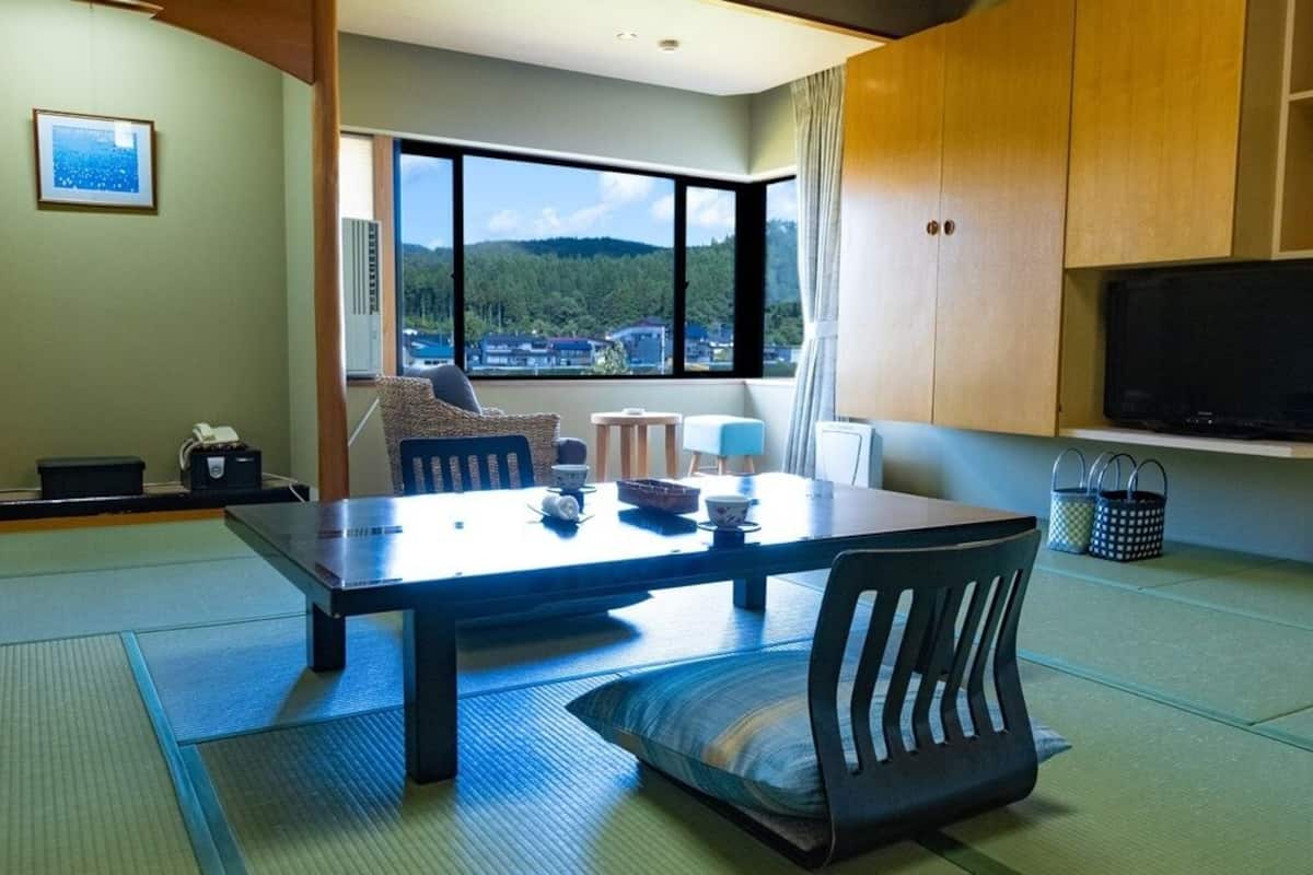 Una habitación con esteras de tatami, una mesa baja con sillas, un televisor y vistas a una zona boscosa.