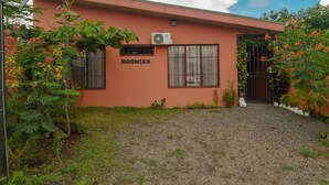 A building labeled "ROOMIES" with a gravel driveway, a small garden with flowering plants, and a clear sky. - Butterfly Apartment (La Fortuna)