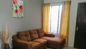 A living room with a brown sectional sofa, a vase with dried plants, a painting of flowers, and a window with yellow curtains. - Butterfly Apartment (La Fortuna)