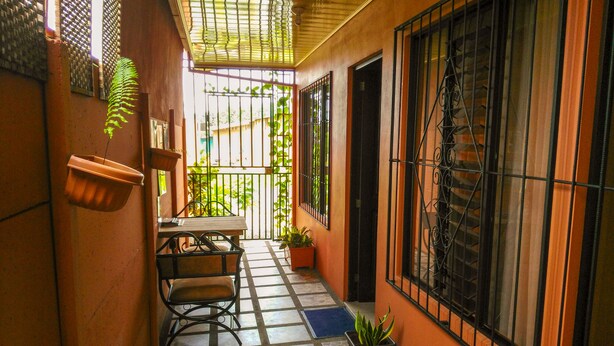 A narrow corridor with a tiled floor, a table with chairs, potted plants, and barred windows. - Butterfly Apartment (La Fortuna)
