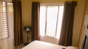 A hotel room with a bed, curtains, a standing fan, and a window with blinds. - Butterfly Apartment (La Fortuna)